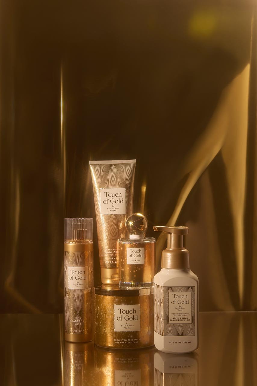 Photo of Touch of Gold collection including hand soap, body cream, 3-wick candle, eau de parfum and fine fragrance mist on a gold background.