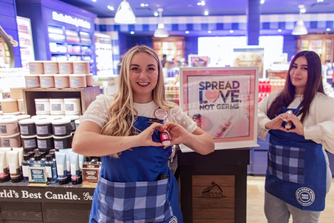Two Bath & Body Works employees in a store, each forming a heart-shape with their hands over a hand sanitizer that is clipped to their apron.