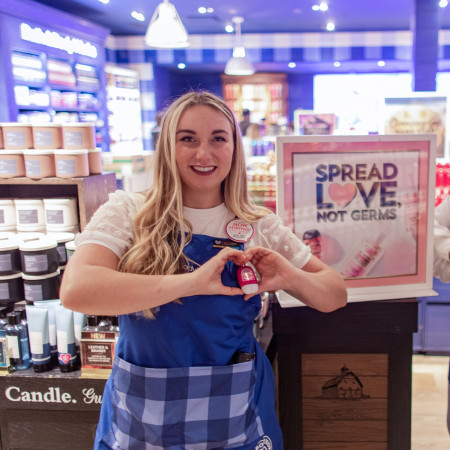 Two Bath & Body Works employees in a store, each forming a heart-shape with their hands over a hand sanitizer that is clipped to their apron.